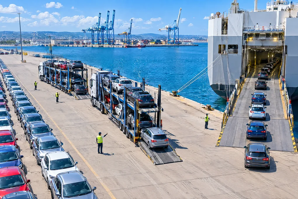 Gebrauchtwagen werden im Hafen einzeln auf ein Autotransportschiff gefahren, daneben entladen offene Autotransporter